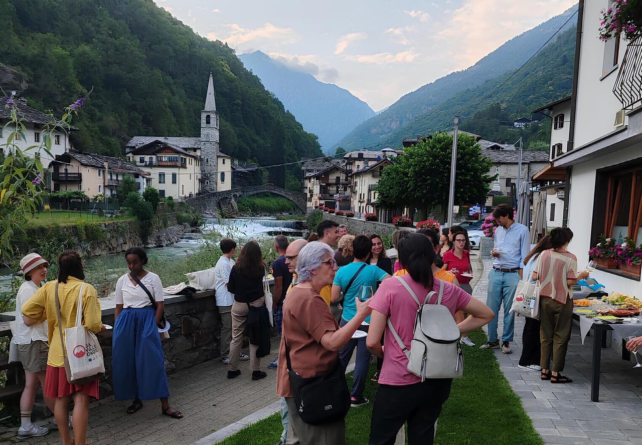 A crowd gathered at a riverside table of food in an alpine village, a stone bell tower and wooded mountains behind