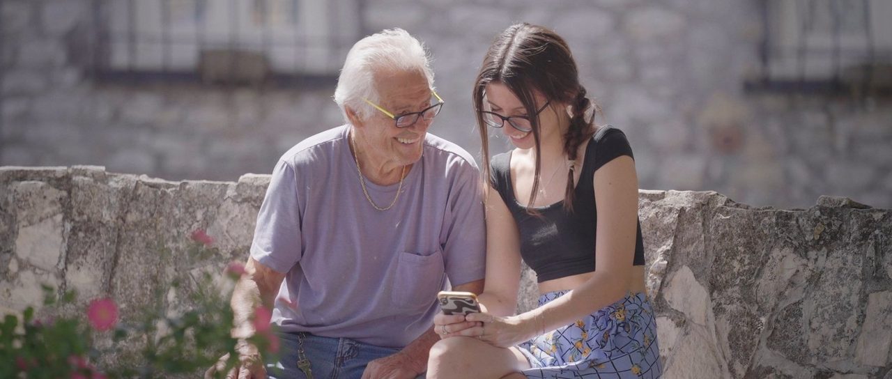 An older man and a young woman looking at a phone together beside a stone wall