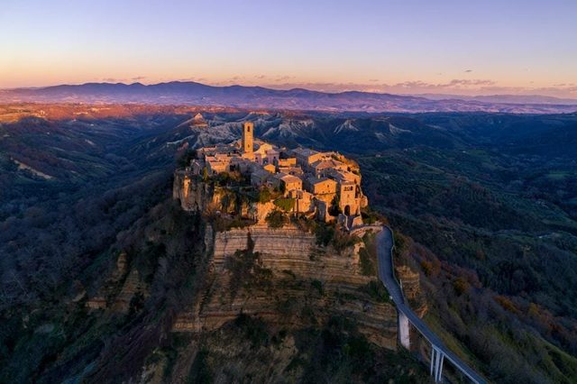 Aerial view at dusk of a hilltop village on an eroded rock outcrop, reached by a long footbridge