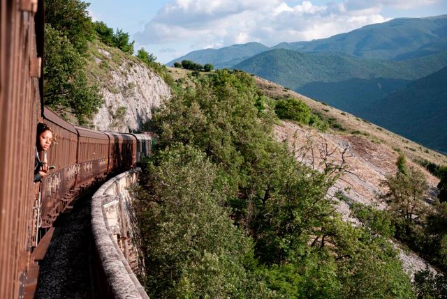 A passenger leaning from the window of a red historic train curving along a stone viaduct through green mountains