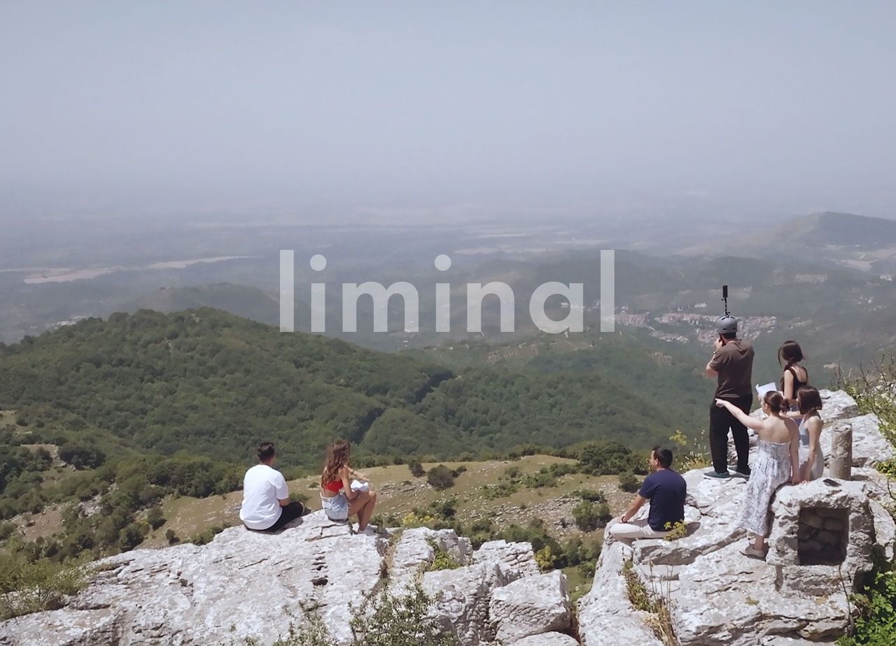People resting on a rocky summit above a hazy wooded plain, the word liminal across the sky