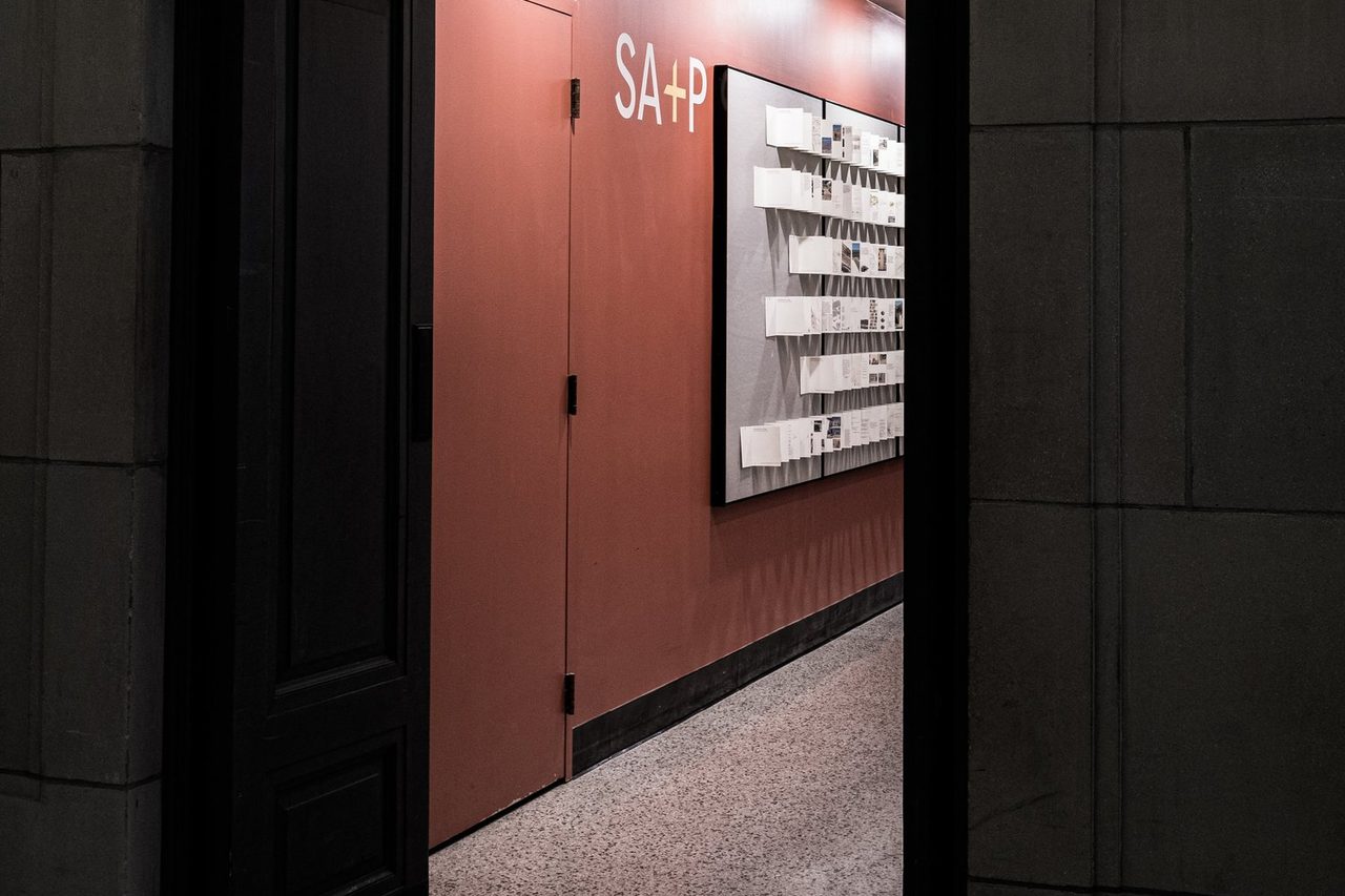 Doorway in a dark stone wall opening onto a terracotta-red corridor hung with exhibition panels