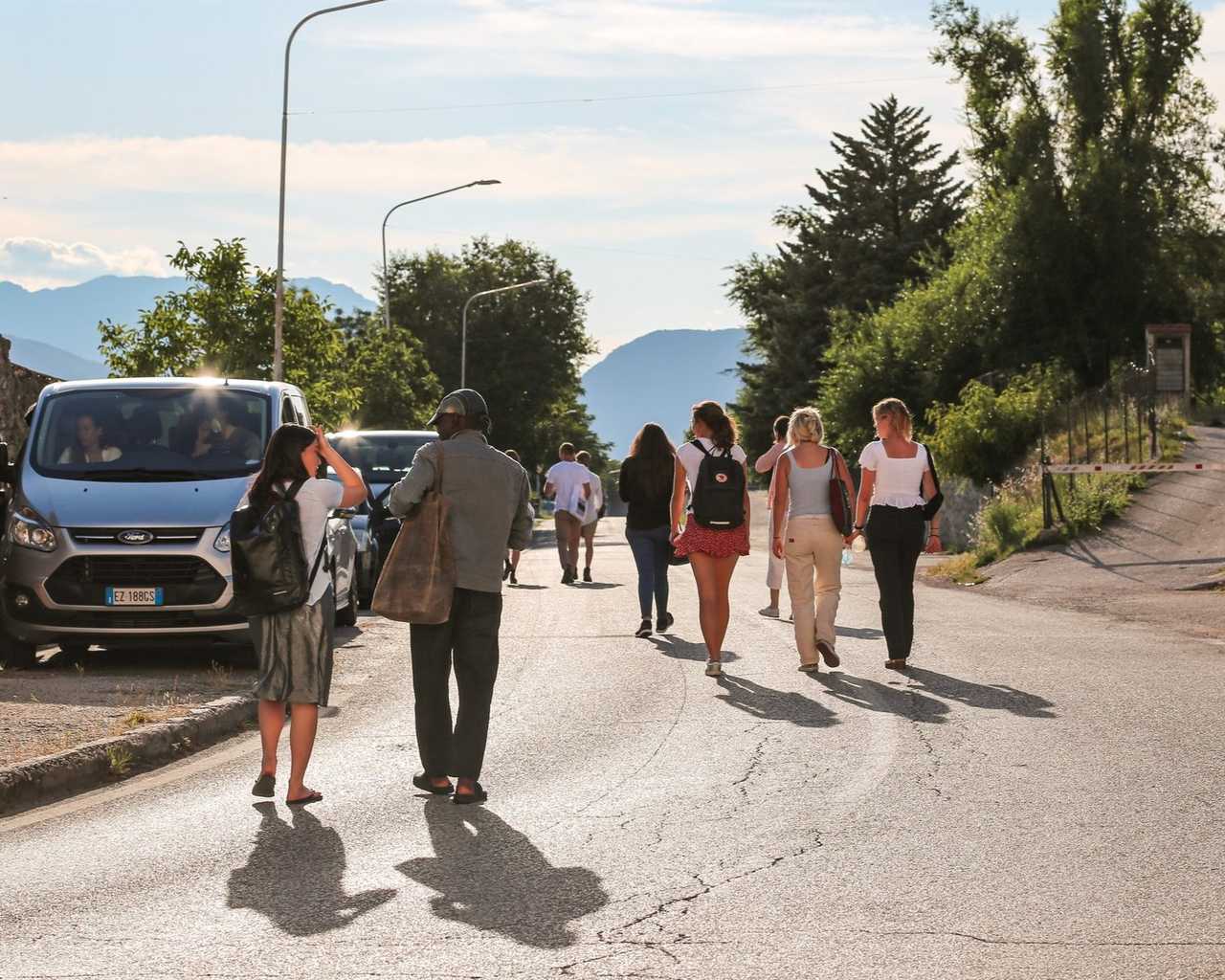 A group walking along a sunlit road lined with trees, hills behind